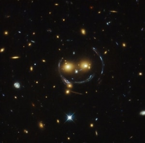 In the centre of this image, taken with the NASA/ESA Hubble Space Telescope, is the galaxy cluster SDSS J1038+4849 — and it seems to be smiling. You can make out its two orange eyes and white button nose. In the case of this “happy face”, the two eyes are very bright galaxies and the misleading smile lines are actually arcs caused by an effect known as strong gravitational lensing. Galaxy clusters are the most massive structures in the Universe and exert such a powerful gravitational pull that they warp the spacetime around them and act as cosmic lenses which can magnify, distort and bend the light behind them. This phenomenon, crucial to many of Hubble’s discoveries, can be explained by Einstein’s theory of general relativity. In this special case of gravitational lensing, a ring  — known as an Einstein Ring  — is produced from this bending of light, a consequence of the exact and symmetrical alignment of the source, lens and observer and resulting in the ring-like structure we see here. Hubble has provided astronomers with the tools to probe these massive galaxies and model their lensing effects, allowing us to peer further into the early Universe than ever before. This object was studied by Hubble’s Wide Field and Planetary Camera 2 (WFPC2) and Wide Field Camera 3 (WFC3) as part of a survey of strong lenses. A version of this image was entered into the Hubble’s Hidden Treasures image processing competition by contestant Judy Schmidt.